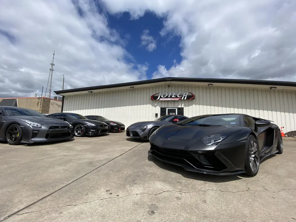 Multiple high-performance cars including a Lamborghini Aventador, Nissan GT-Rs, and a Toyota Supra parked outside Jotech Motorsports.