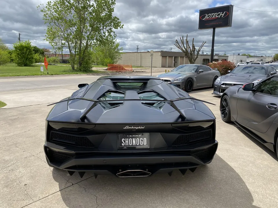 Rear view of a matte black Lamborghini Aventador parked at Jotech Motorsports with Nissan GT-Rs and a Toyota Supra.