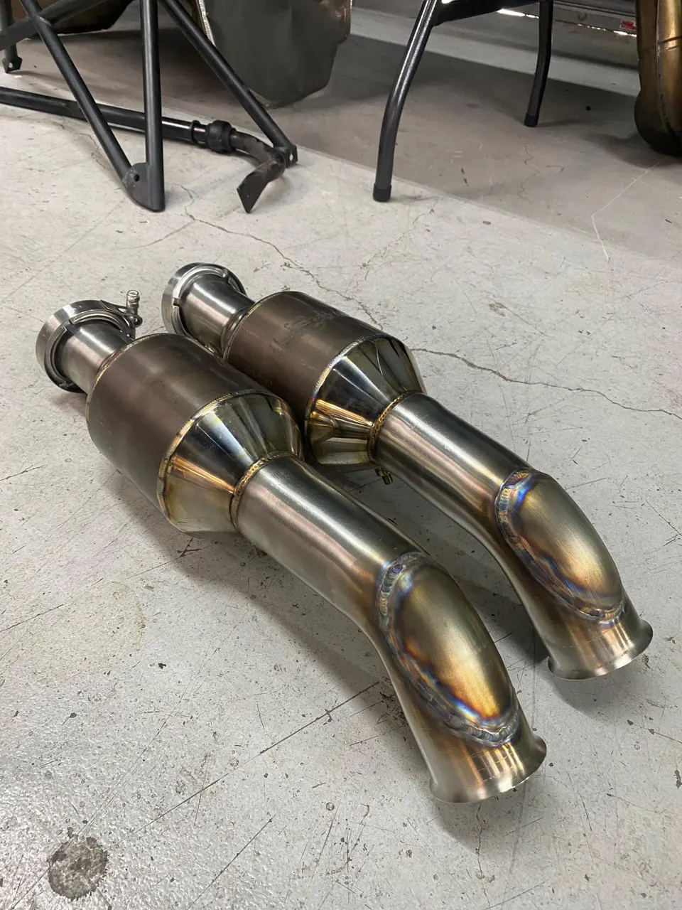 Two stainless steel exhaust downpipes with V-band clamps and custom welds on a concrete shop floor.