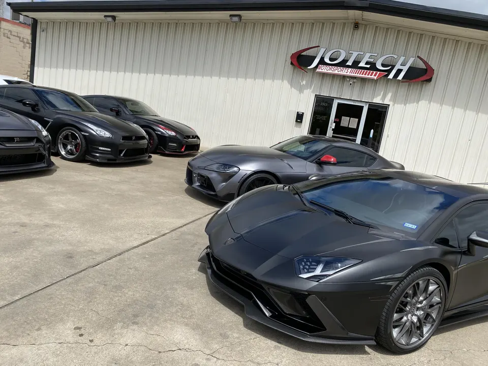 A group of high-performance sports cars including Nissan GT-Rs, a Toyota Supra, and a Lamborghini Aventador parked outside Jotech Motorsports.