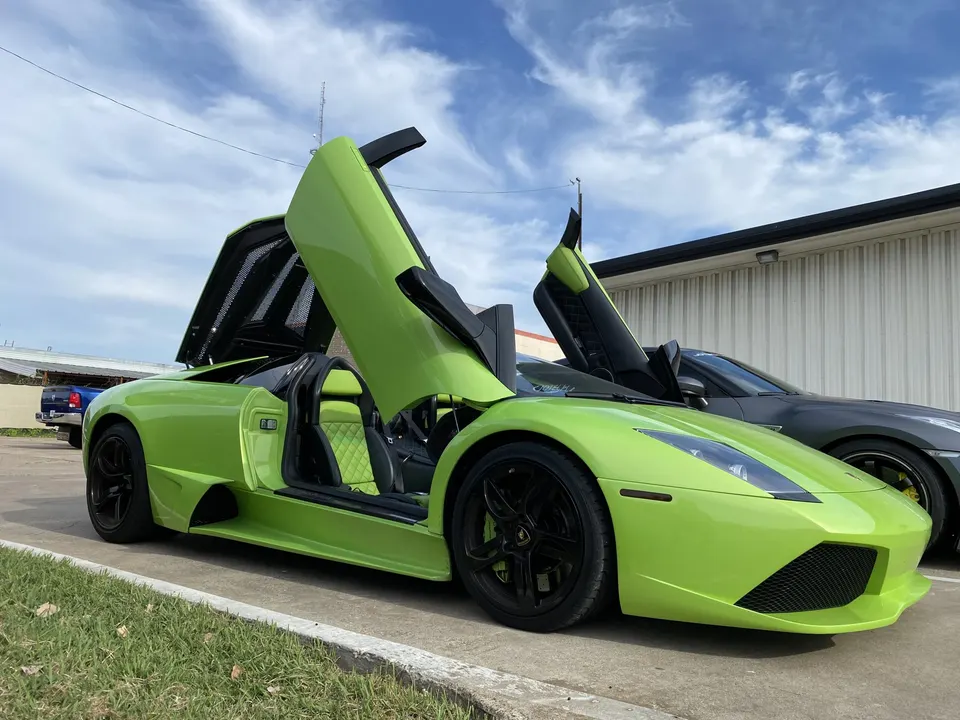 Lime green Lamborghini Murcielago with scissor doors open, parked outdoors next to another sports car.