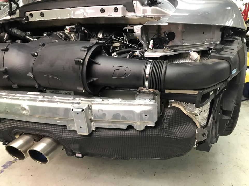 Rear view of a silver Porsche with the bumper removed, showcasing a performance intake system and exhaust tips during an install.