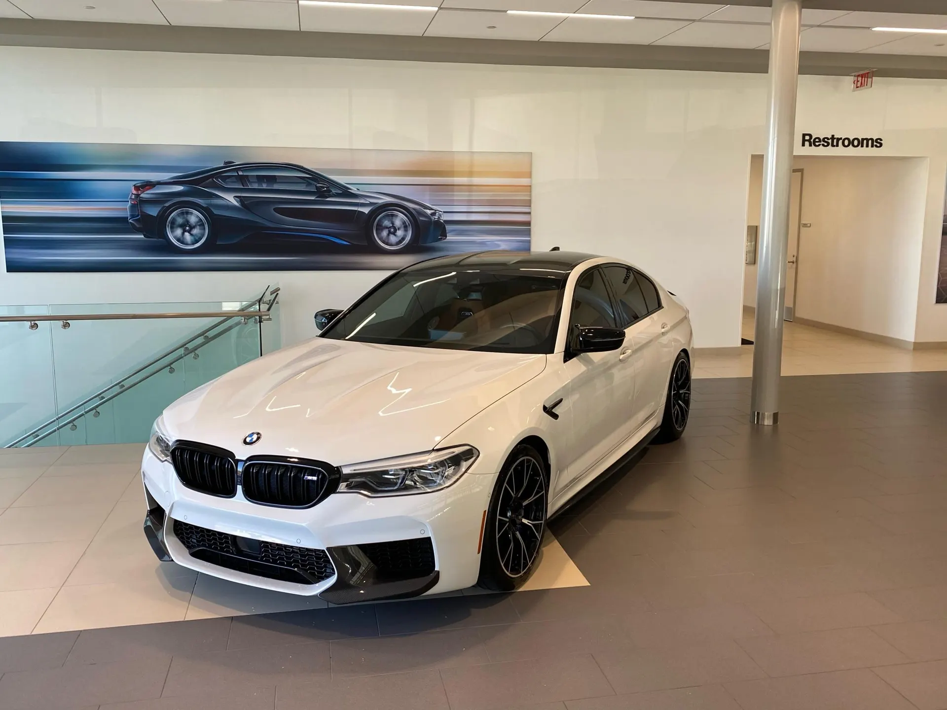 A white BMW M5 luxury sedan displayed in a modern car dealership showroom with a large car graphic on the wall.