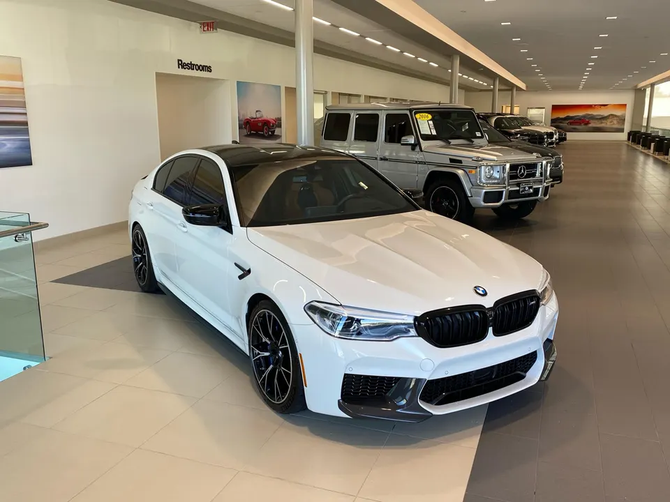 White BMW M5 sedan on display in a car dealership showroom with other luxury vehicles including a Mercedes G-Wagen.