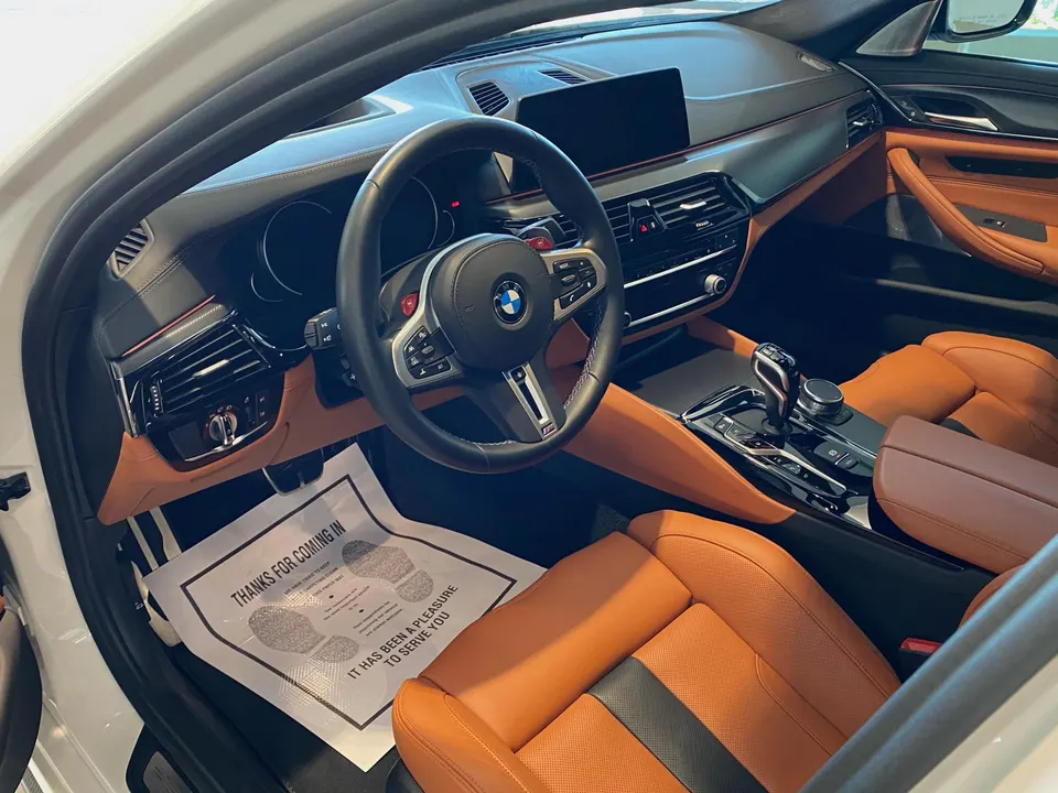 Interior view of a white BMW M5 with brown leather seats, steering wheel, and dashboard, featuring a 'Thanks for Coming In' floor mat.