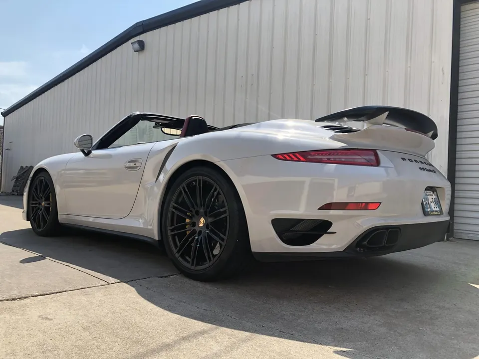 White Porsche 911 Turbo S convertible with black wheels and a rear wing parked outside a building.