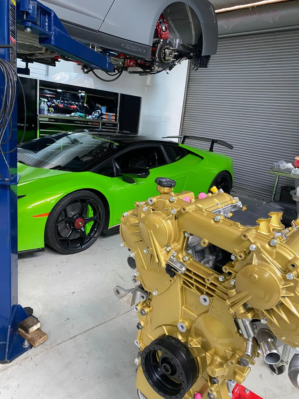 A bright green Lamborghini Huracan in a performance shop with a gold-painted engine block in the foreground and a grey car on a lift.