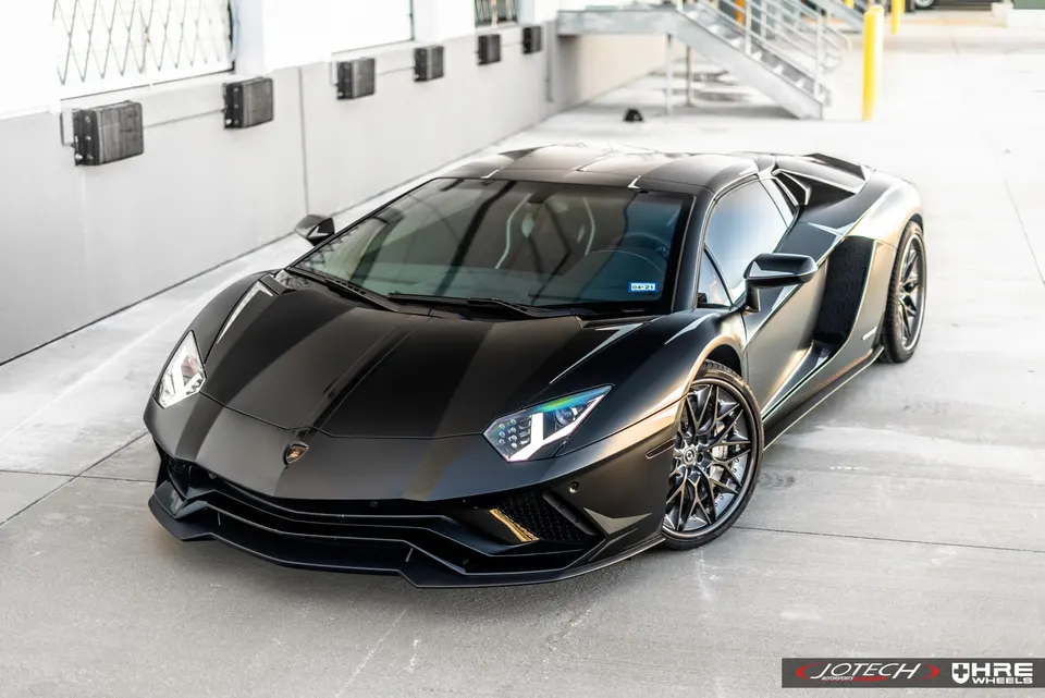 A black Lamborghini Aventador with HRE wheels parked on a concrete lot, showcasing its sleek design.