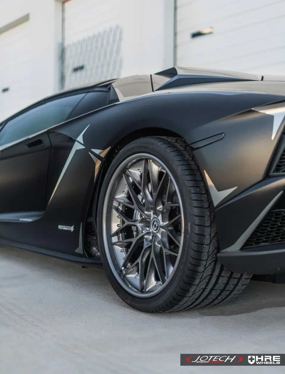 Matte black Lamborghini Aventador with custom HRE wheels and performance tires, parked in front of a garage.