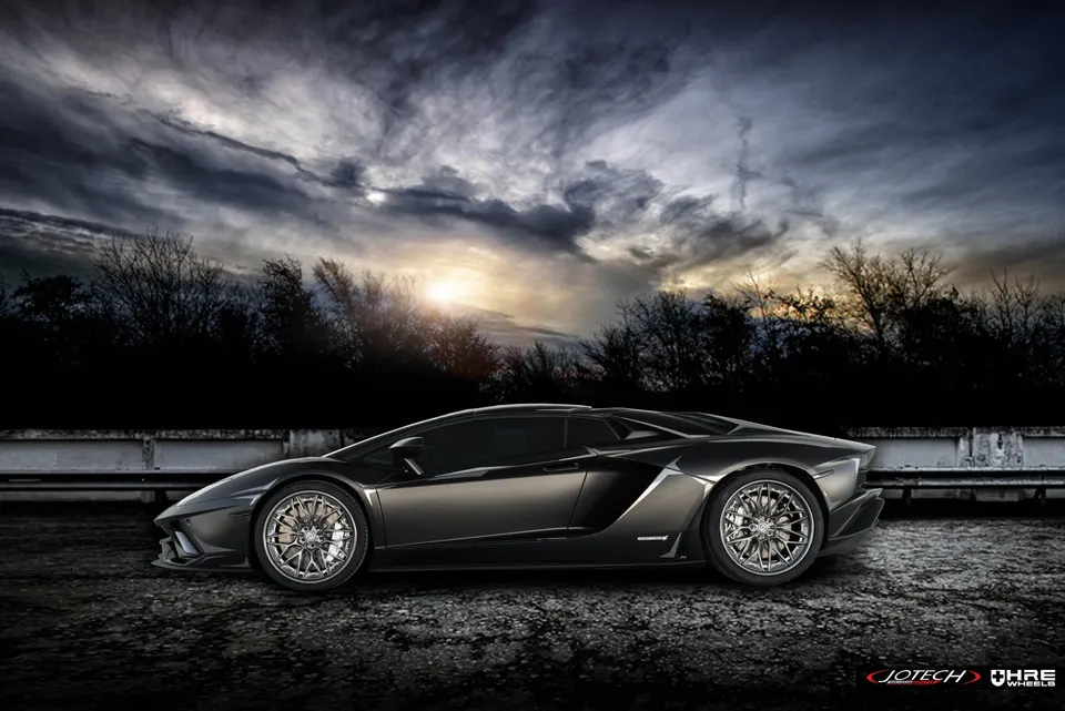 Dark grey Lamborghini Aventador with HRE aftermarket wheels parked under a dramatic cloudy sky at sunset.