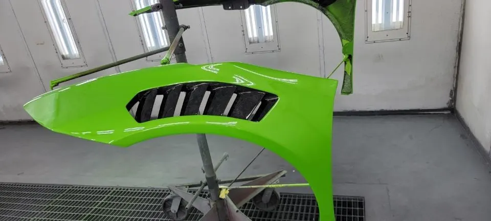 Bright green car fender with a black vent hanging on a stand in a paint booth at an automotive shop.