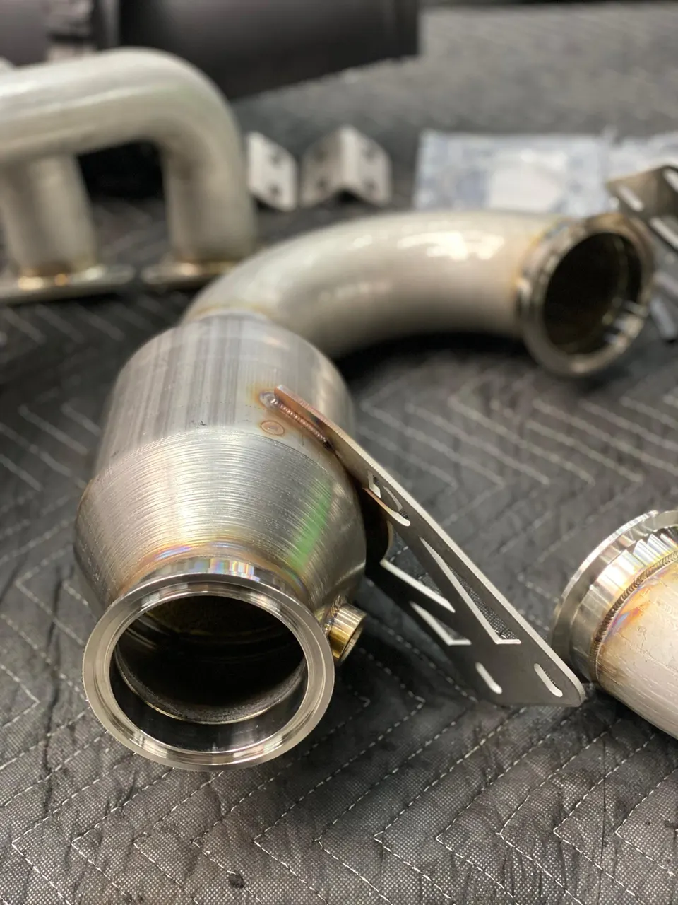 Close-up of new stainless steel exhaust components, including a catalytic converter and piping, on a shop mat.