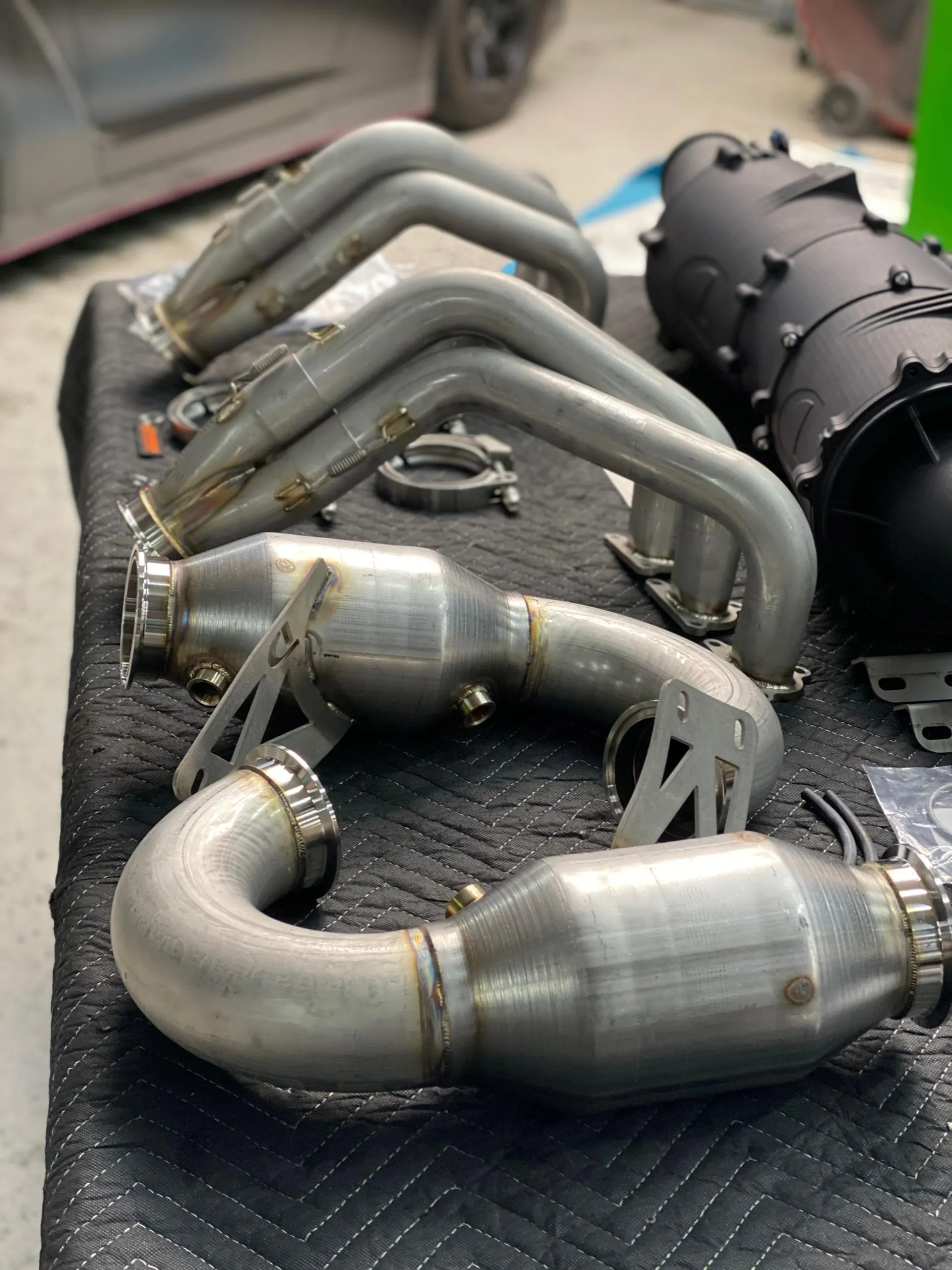 Stainless steel exhaust headers and catalytic converters laid out on a table in a performance tuning shop.