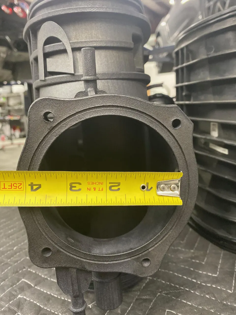 A close-up of a large diameter intake manifold or throttle body being measured with a yellow tape measure in a shop.