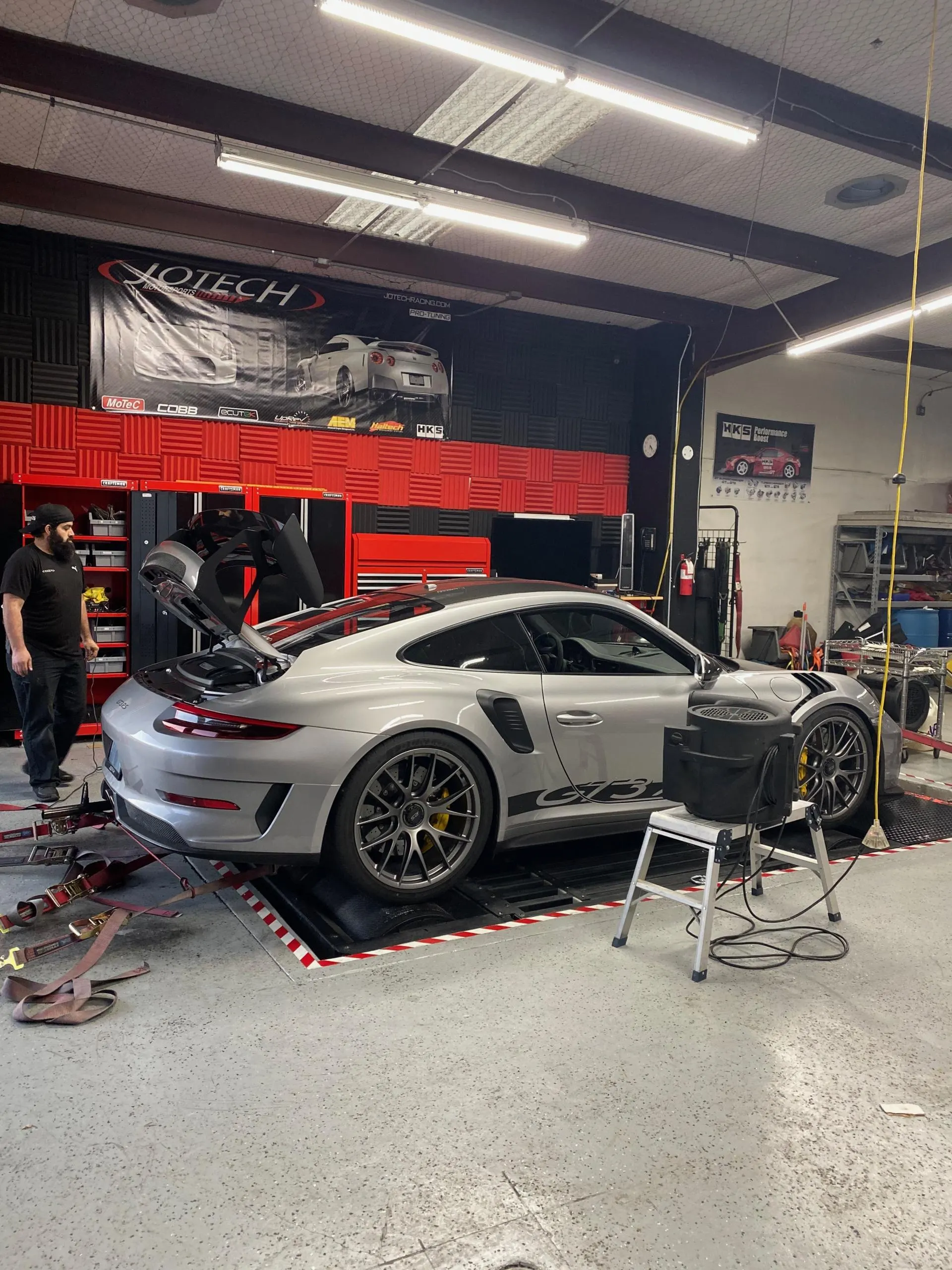 A silver Porsche GT3RS strapped to a dynamometer at Jotech Motorsports for performance tuning.