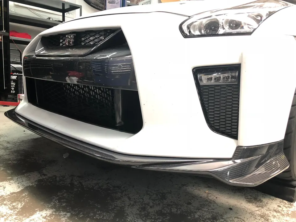 White Nissan GT-R with a carbon fiber front splitter and aero elements installed on the front bumper in a shop.