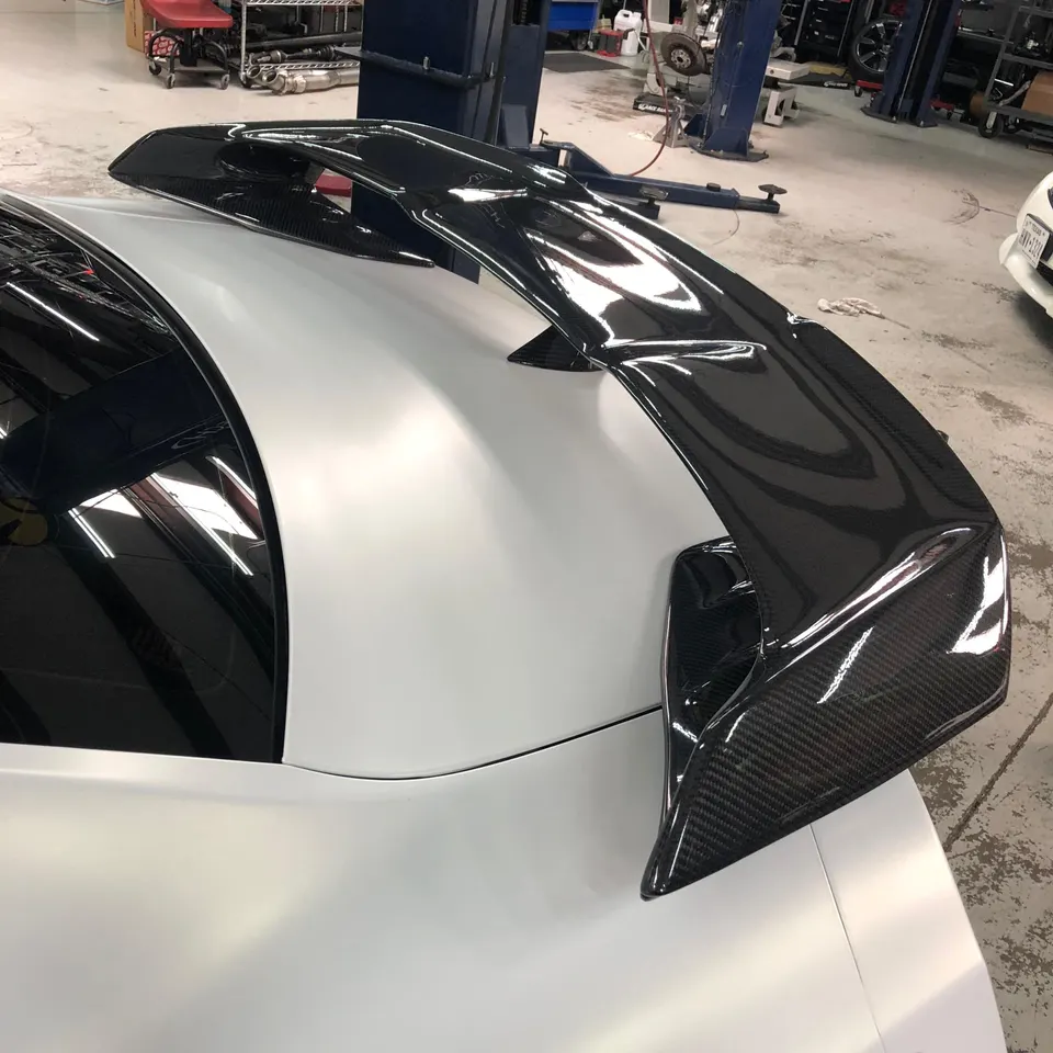 Close-up of a glossy carbon fiber rear wing installed on a white sports car in a performance shop.