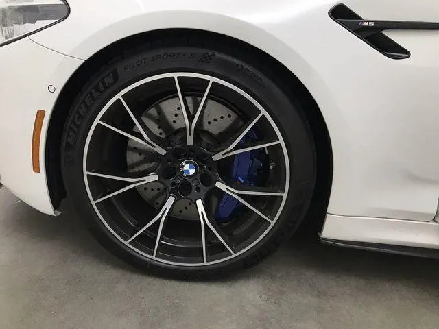A close-up of a white BMW M5's front wheel, showing the black multi-spoke rim, Michelin tire, and blue brake caliper.