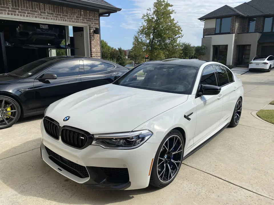 White BMW M5 parked in a driveway with a black Tesla Model S and a white Tesla Model X in the background.