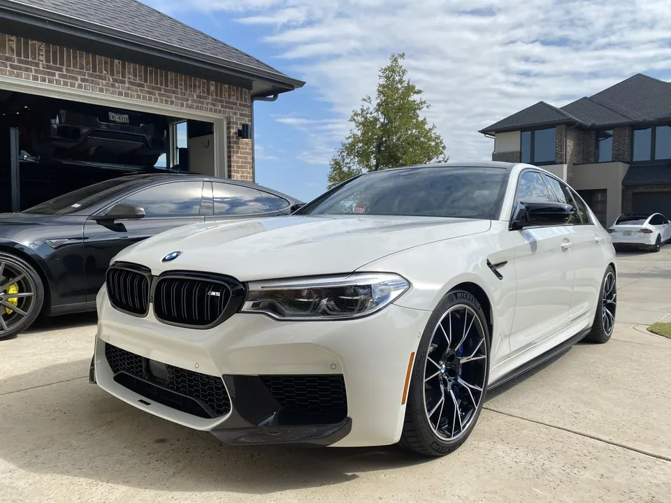 White BMW M5 performance sedan parked in a residential driveway with a black Tesla Model S and another car in the garage.