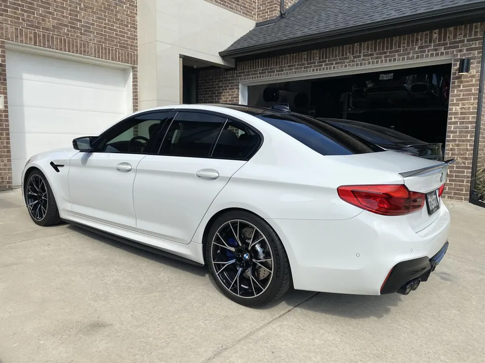 White BMW M5 sedan parked in a driveway in front of a brick house with an open garage.