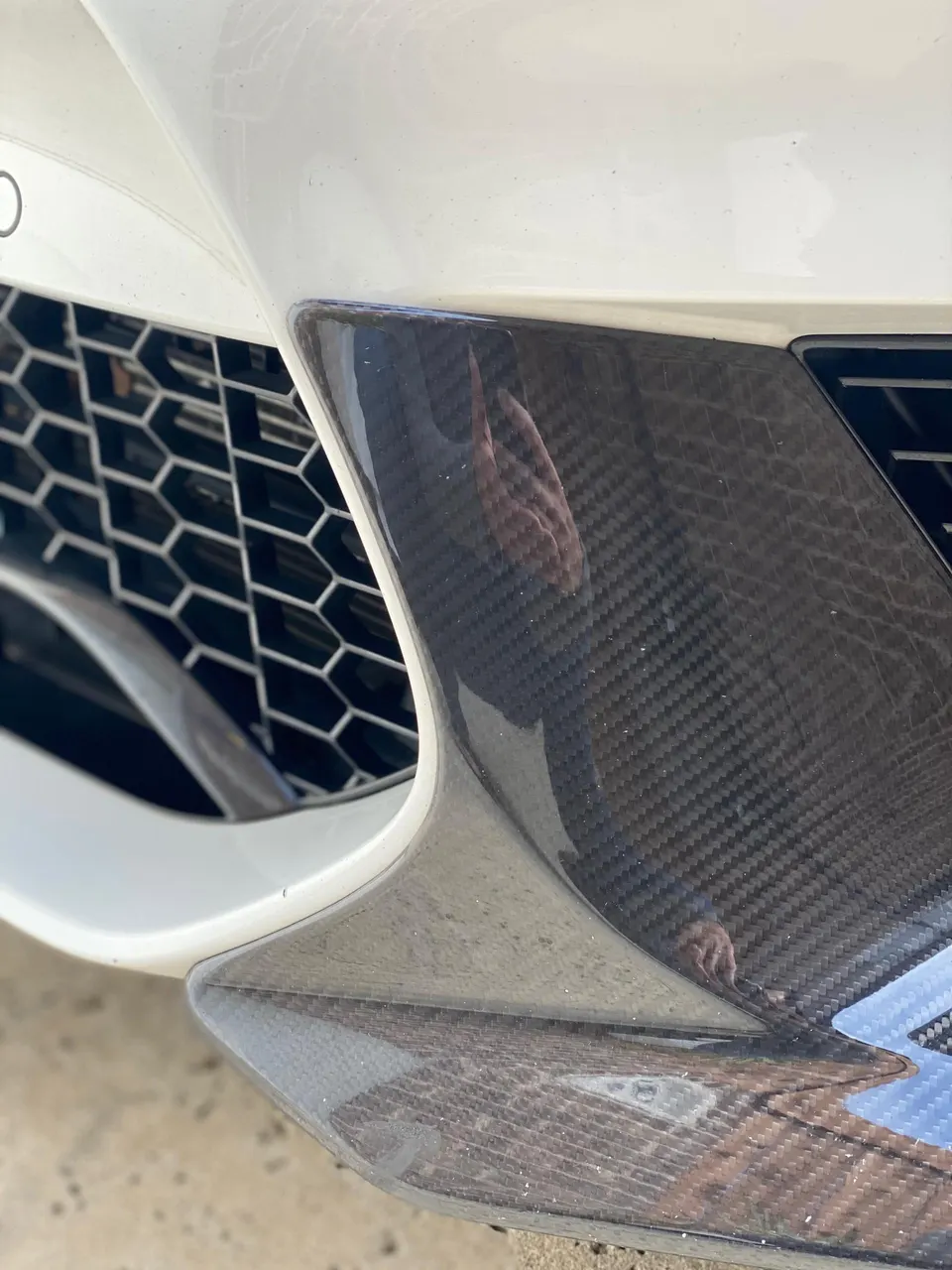 A close-up of a white car's front bumper featuring a honeycomb grille and carbon fiber aerodynamic trim.