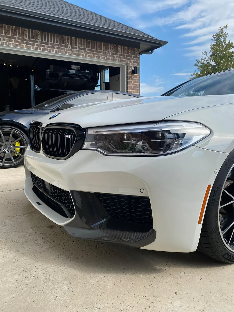 White BMW M5 luxury sedan with a carbon fiber front splitter parked outside a garage with another car on a lift.