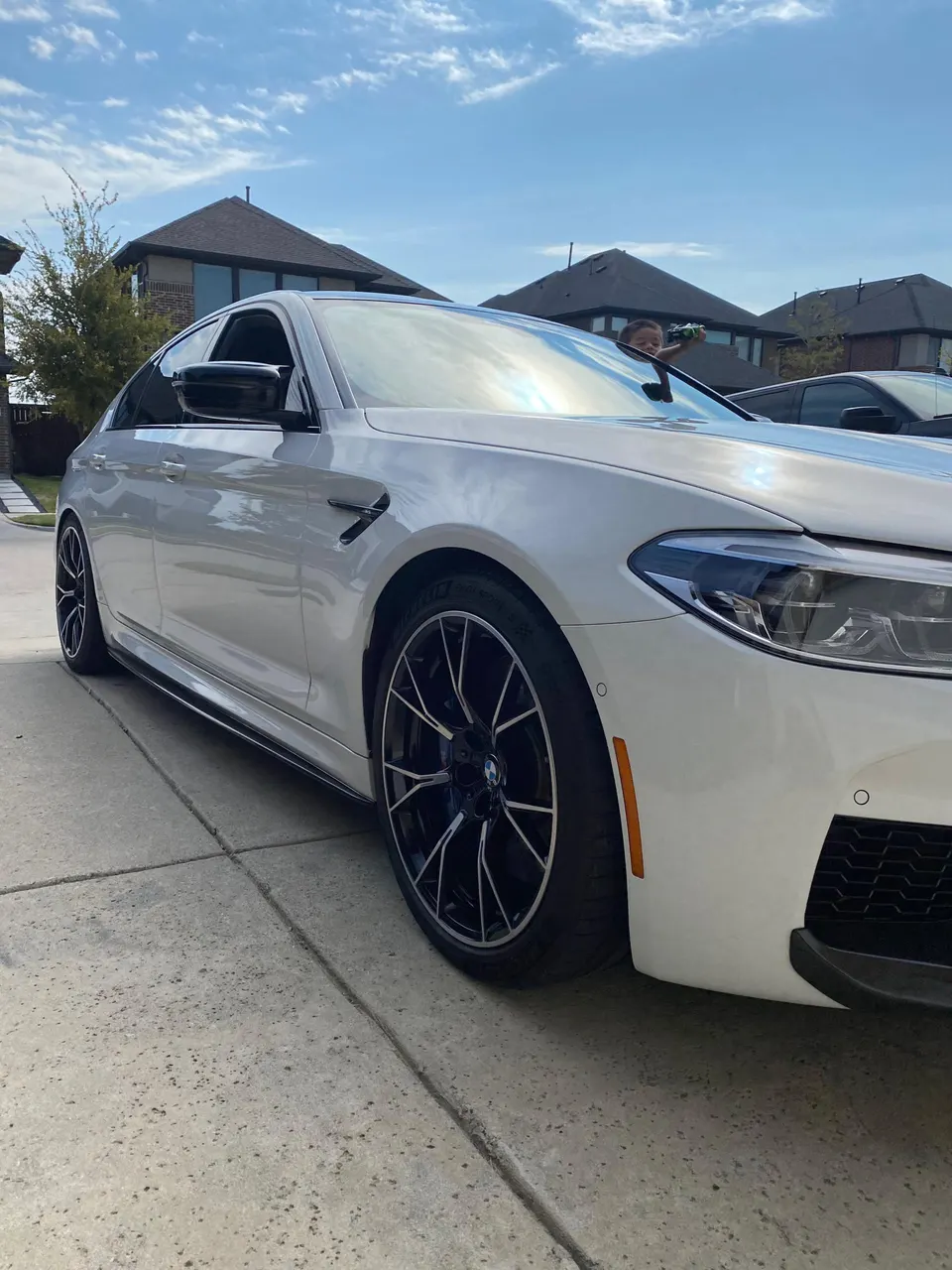 White BMW M5 parked on a residential driveway, showcasing aftermarket black wheels and a side skirt.