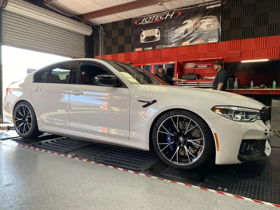 A white BMW M5 sedan is strapped onto a dynamometer at Jotech Motorsports, with technicians nearby in the shop.
