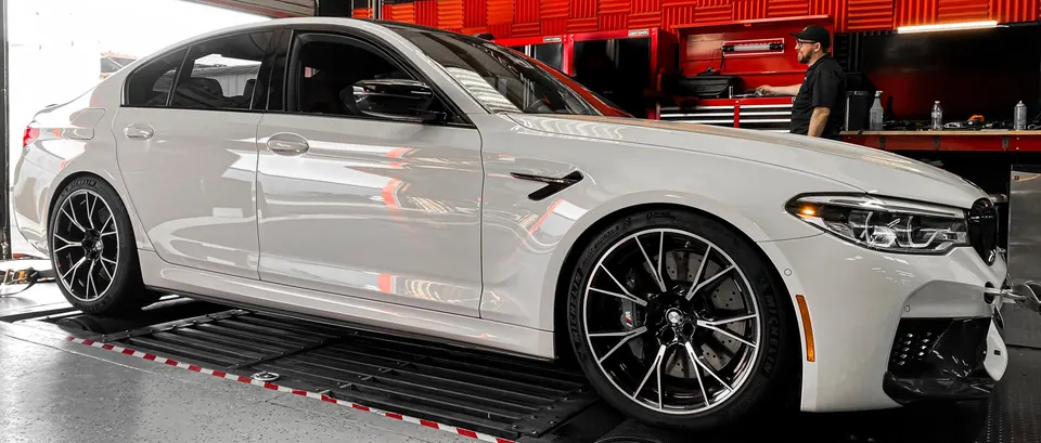 A white BMW M5 sedan is strapped onto a dynamometer in a performance tuning shop, with a technician in the background.