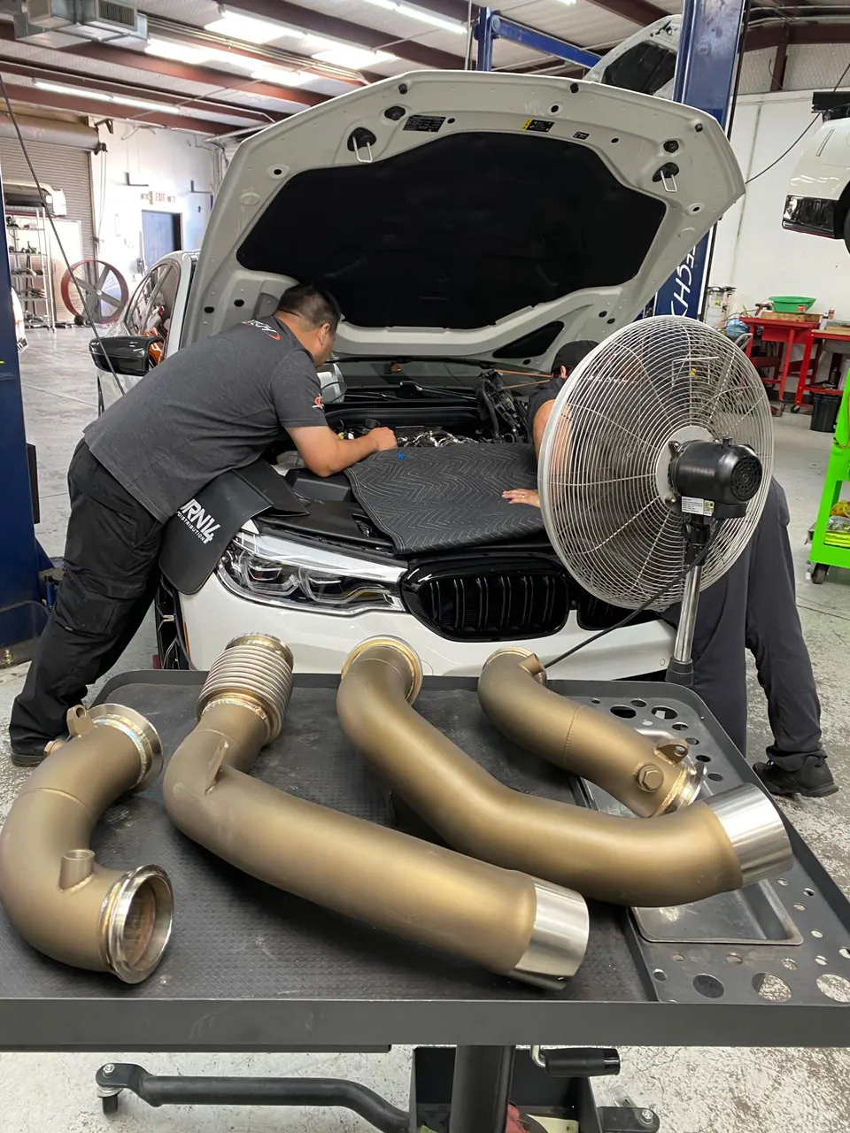 Mechanics installing aftermarket exhaust downpipes on a white BMW M series in a performance tuning shop.