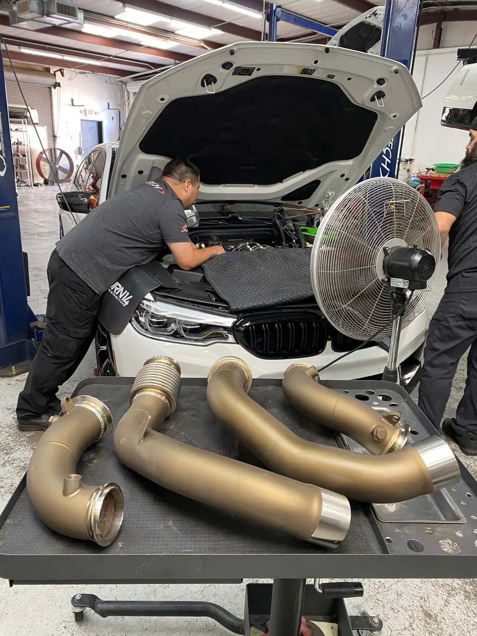 A mechanic works on a white BMW M car on a lift in a shop, with aftermarket exhaust downpipes on a table.