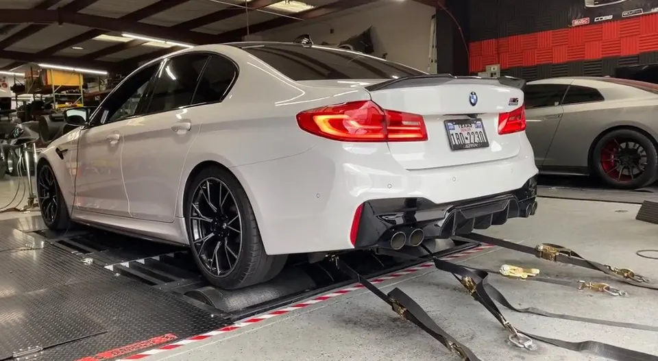 White BMW M5 sedan strapped to a dynamometer for performance testing at a tuning shop, with a Nissan GT-R in the background.