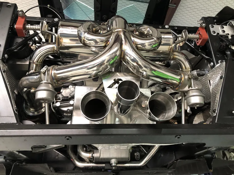 A detailed overhead view of a polished aftermarket performance exhaust system on a high-performance car, likely a Lamborghini.