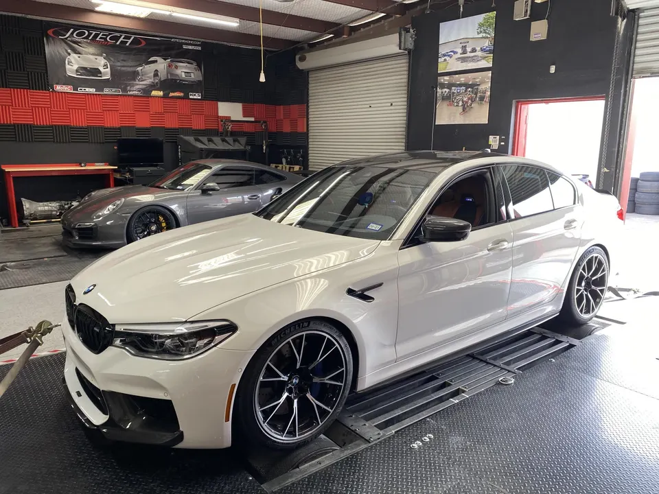 White BMW M5 sedan on a dynamometer at Jotech Motorsports, with a grey Porsche 911 in the background.