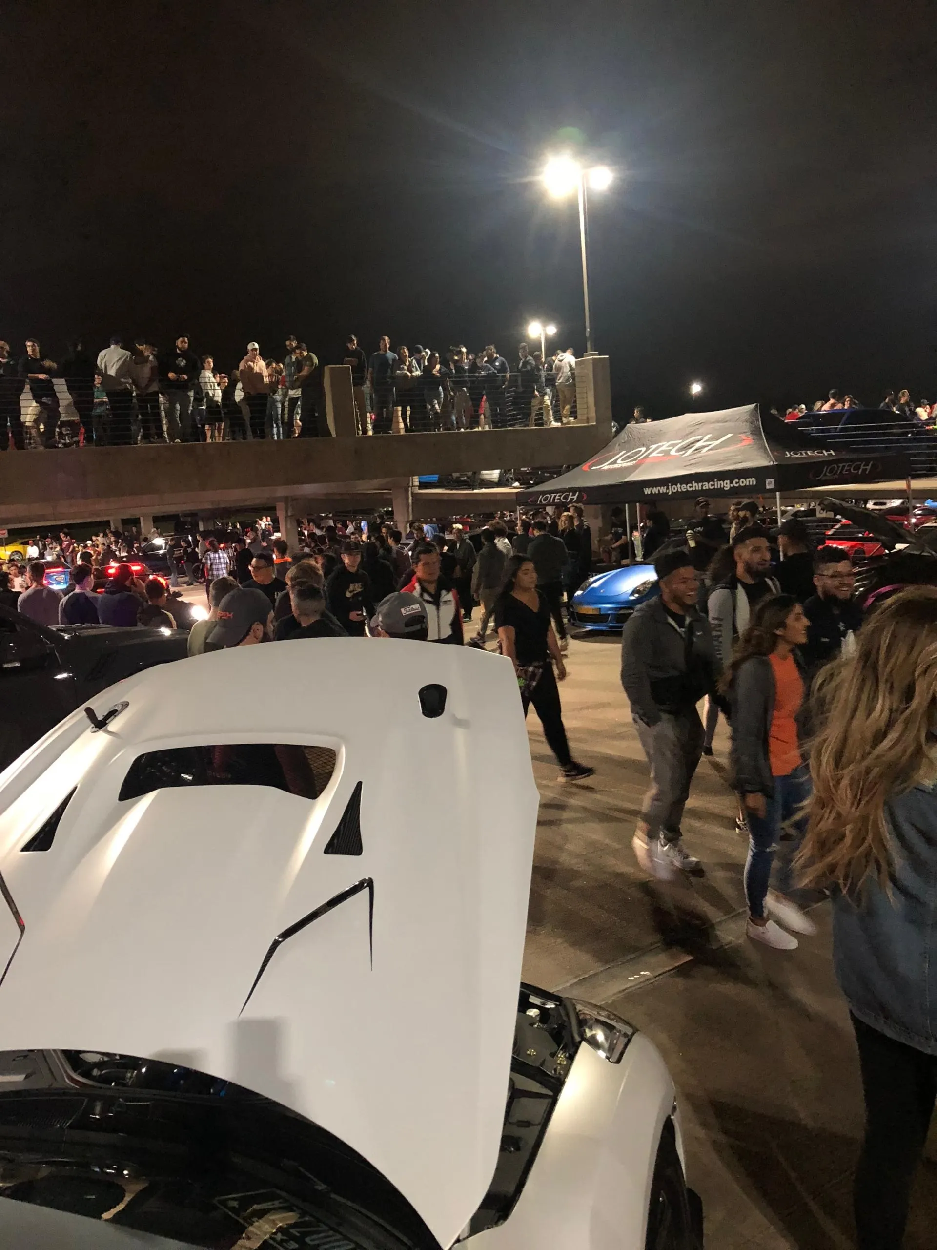Crowd gathered at a night car meet with a white performance car in the foreground and a Jotech tent in the background.