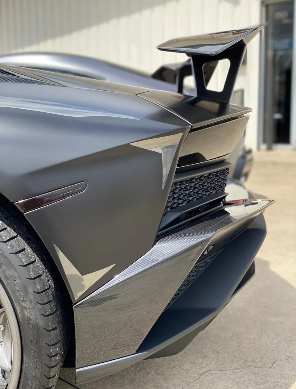 Close-up of the matte black rear of a Lamborghini supercar, showcasing its carbon fiber wing and diffuser.