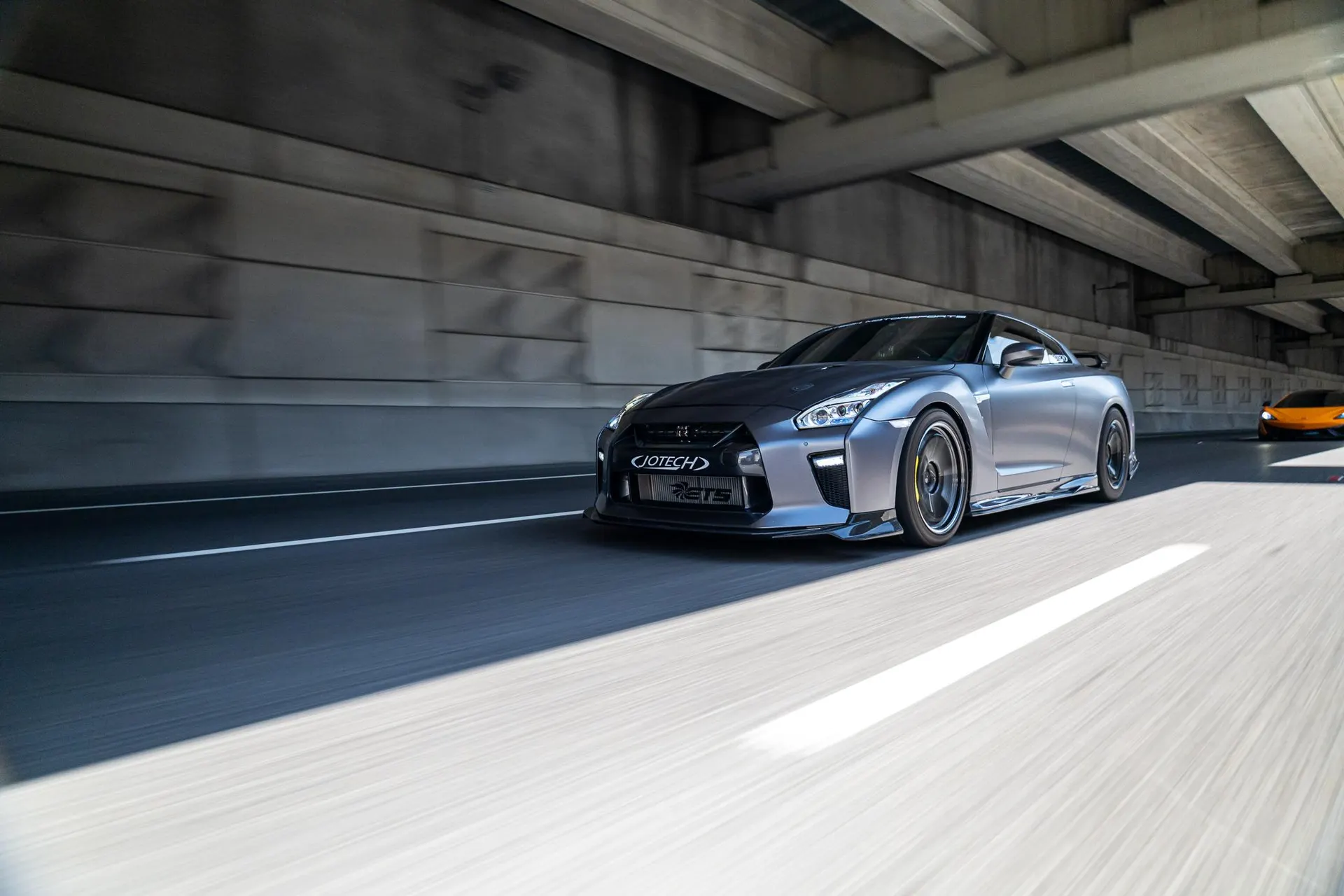 A grey Nissan GT-R with Jotech and ETS branding drives on a highway with an orange McLaren in the background.