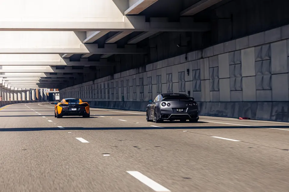Orange McLaren and grey Nissan GT-R driving side-by-side on a multi-lane highway underpass.
