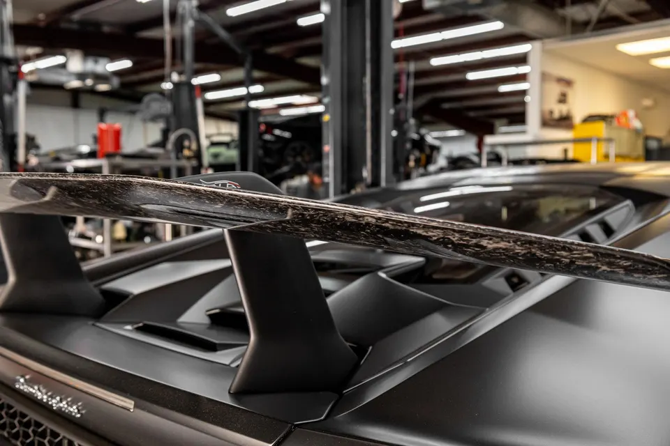 Close-up of a matte black Lamborghini with a carbon fiber rear wing in a performance shop.