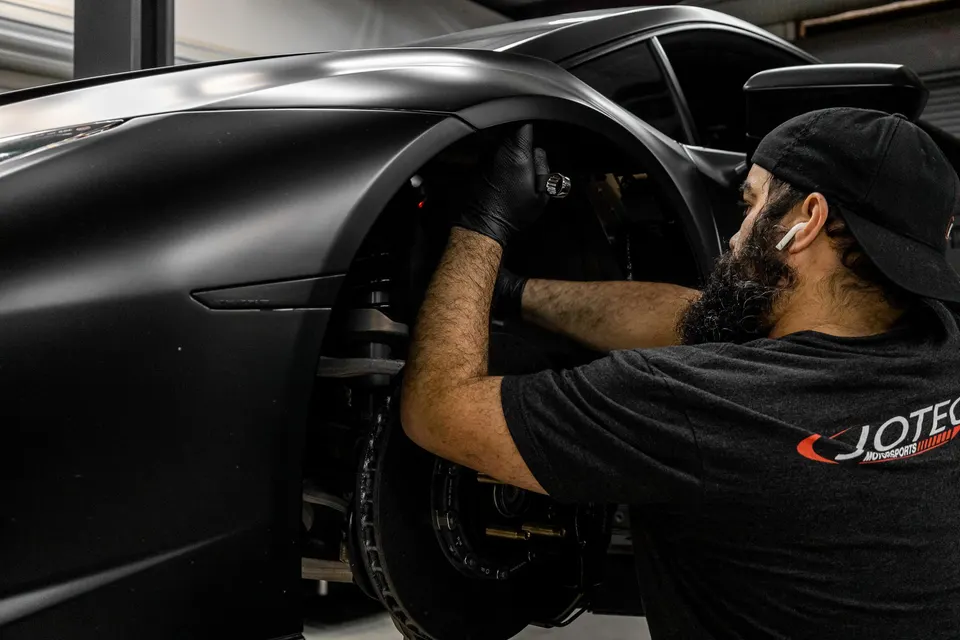 A mechanic in black gloves works on the front wheel and suspension of a matte black Lamborghini Huracan in a performance shop.