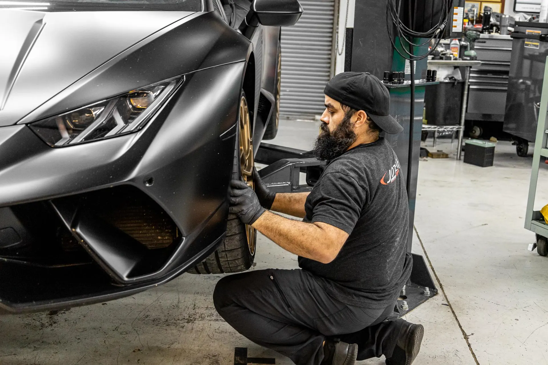 Mechanic working on the front wheel of a matte black Lamborghini Huracan on a lift in a performance tuning shop.
