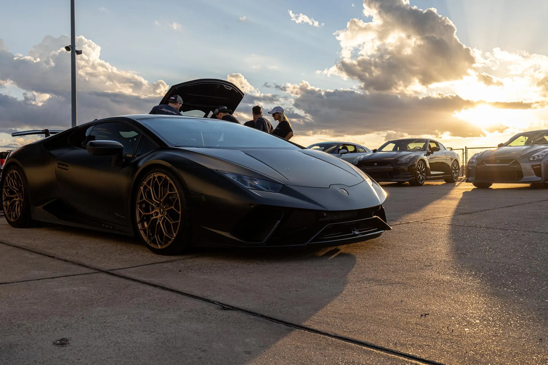 Matte black Lamborghini Huracan and Nissan GT-Rs parked at a car meet during sunset with people gathered around.
