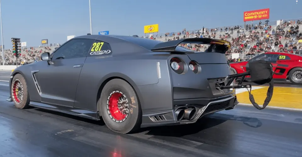 A grey Nissan GT-R with drag racing modifications, including a parachute and drag wheels, on a drag strip.