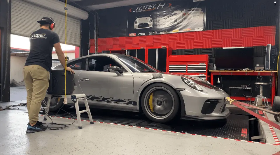 A silver Porsche 911 GT3 RS strapped to a dyno at Jotech Motorsports, with a technician operating equipment.