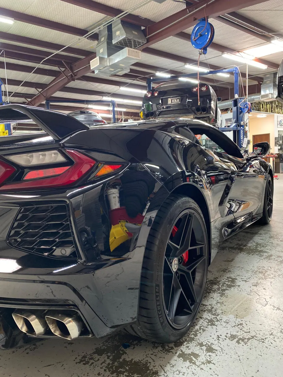 Rear view of a black Chevrolet Corvette C8 with aftermarket exhaust tips and HRE wheels in a performance tuning shop.