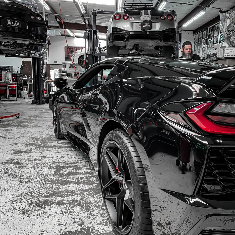 A black Corvette C8 in a performance tuning shop with a silver Nissan GT-R on a lift in the background.
