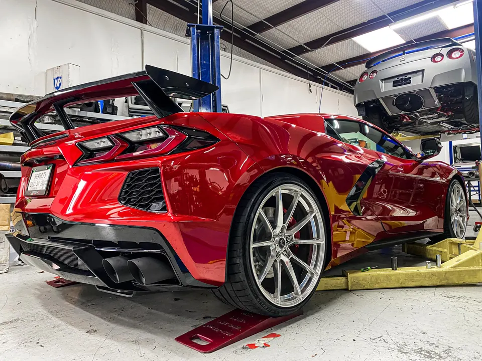 A red Corvette C8 with an aftermarket wing and custom wheels on a lift in a performance shop, with a Nissan GT-R on another lift.