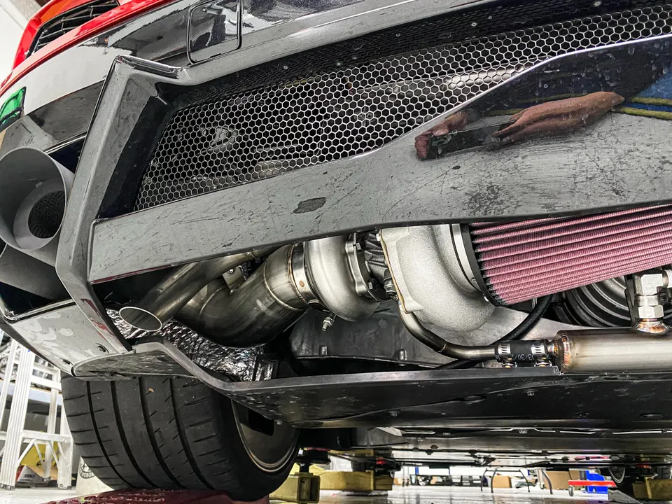 Rear underside view of a Lamborghini with a large turbocharger, performance exhaust, and air filter installed in a shop.