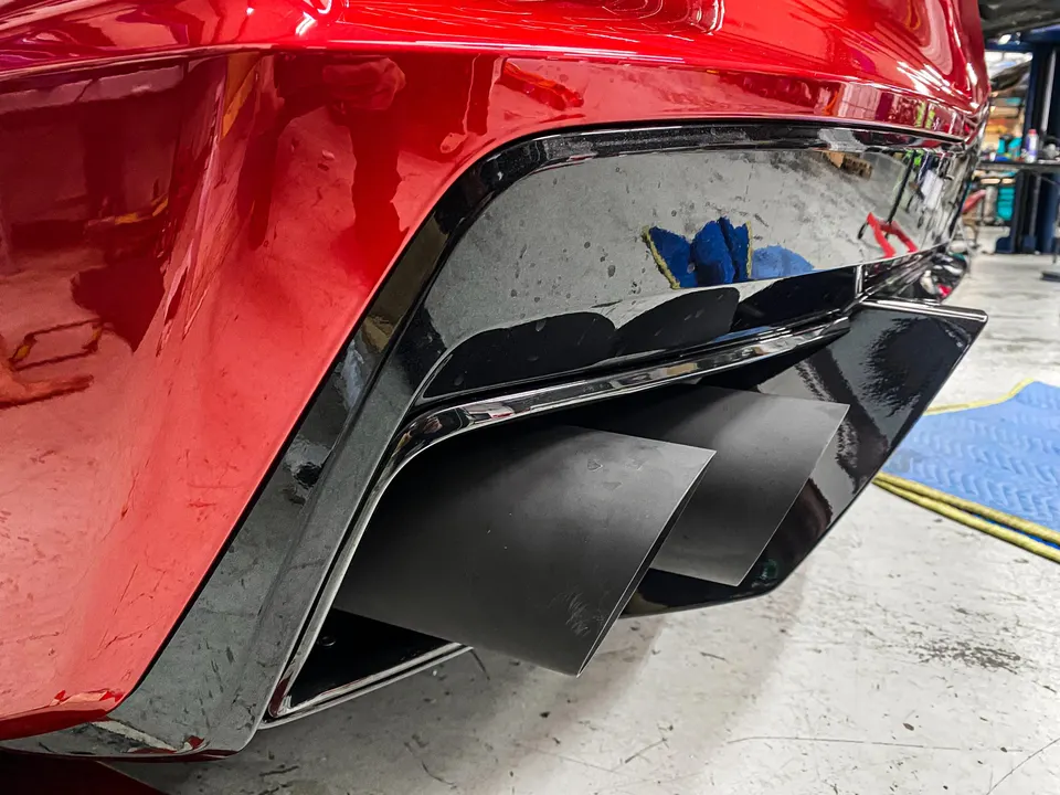 Close-up of a red car's rear bumper with black aftermarket exhaust tips in a shop setting.